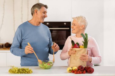 Market alışverişi. Neşeli, yaşlı bir çift, evdeki modern mutfaktaki sebze ve sebzelerle dolu paketle yemek pişiriyor. Kıdemli eşler evde birlikte yemek hazırlıyor. Sağlıklı Beslenme