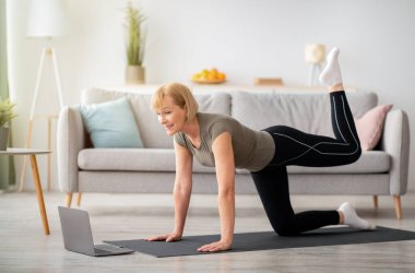 Çevrimiçi fitness konsepti. Oturma odasında dizüstü bilgisayarla spor videosu izleyen güzel, olgun bir kadın. Formda kıdemli bayan idman yapıyor, kapalı alanda iyi durumda.