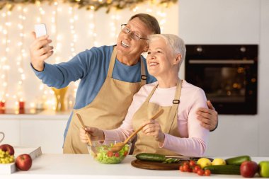 Akıllı telefondan selfie çeken yaşlı bir çift modern mutfaktaki Noel 'i kutlarken eğleniyor. Kıdemli eşler Sağlıklı Noel Yemeği Konseptinin tadını çıkarıyorlar. Seçici Odaklanma