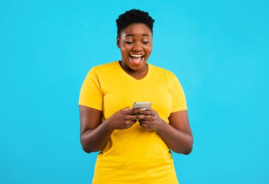 Heyecanlı Afrikalı Büyük Boy Kadın Telefon Uygulaması Mavi Stüdyo Arkaplanının Üzerinde Duruyor. Siyah kadın cep telefonuyla cep telefonu oyunu oynuyor. Akıllı telefon kavramın için harika bir uygulama.