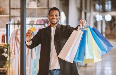 Kolay ödemeler ve mevsimlik satışlar. Binlerce yıllık neşeli Afro-Amerikan adam kredi kartıyla poz veriyor ve alışveriş merkezinde renkli çantalar alıyor, modern mağaza vitrininin yanında alışveriş yapıyor, uzay kopyalarını kopyalıyor.