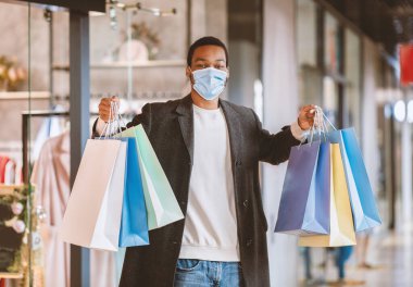 Covid-19 salgınında ve sosyal mesafede alışveriş ve Kara Cuma. Koruyucu maskeli ve ceketli mutlu Afro-Amerikan genç adam modern şehir alışveriş merkezinde bir sürü renkli kese kağıdı gösteriyor.