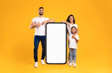 Stüdyoda Sarı Arkaplan 'ın üzerinde duran boş ekranlı mobil reklam uygulamalı büyük akıllı telefonun yanında duran Arap aile. Harika Uygulama Reklamı. İnsanlar ve Gadget Model