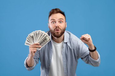 Heyecanlı milenyum adamı elinde para tutuyor, mavi stüdyo arka planında büyük bir galibiyete karşı EVET işareti yapıyor. Dolarlık parası olan genç bir adam finansal başarı elde ediyor, kumarhane bahsini kazanıyor.