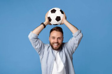 Mavi stüdyo arka planında futbol topunu başının üstünde tutan gülümseyen genç adam. Şampiyonluk maçında en sevdiği takımı için tezahürat yapan pozitif futbol taraftarı, spor maçındaki zaferi kutluyor.