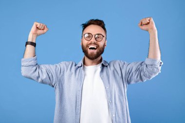 Gözlüklü aşırı neşeli sakallı adam iki eli mavi stüdyo arka planında EVET hareketi yapıyor, panorama. Başarı ya da başarıyı kutlayan muzaffer genç adam, büyük başarı ya da büyük şans.