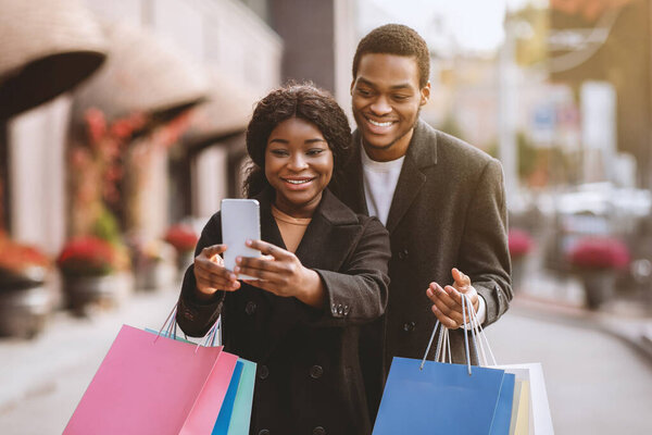 Веселитесь вместе и селфи во время Черной пятницы и распродажи сезона. Улыбающиеся африканские американец тысячелетия и женщина с цветными сумками делают селфи на открытом воздухе, рядом с торговым центром для социальных сетей