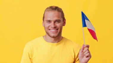 Açık renk saçlı neşeli genç bir adam parlak bir şekilde gülümserken küçük bir Fransız bayrağı tutuyor. Parlak sarı bir arka plana karşı duruyor, pozitif enerji yayıyor ve kutlama yapıyor..