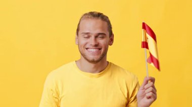 Afrika ve Kafkas özellikleri olan neşeli bir genç adam elinde küçük bir İspanyol bayrağı tutarken gülümsüyor. Parlak sarı bir gömlek giyer, parlak sarı bir arkaplanda durur..