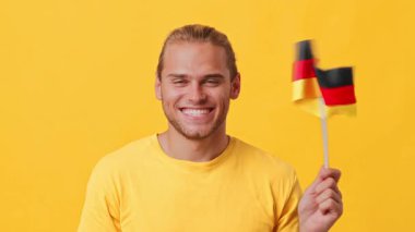Parlak bir gülümsemesi olan genç bir adam elinde Alman bayrağı gösteriyor. Sarı bir gömlek giyiyor ve canlı sarı bir arka planın önünde duruyor. Ortam neşeli ve canlı..