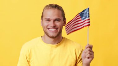 Miras sahibi genç bir adam Amerikan bayrağını tutuyor ve coşkulu bir şekilde gülümsüyor. O sıradan sarı bir gömlek giyiyor ve canlı sarı bir arkaplanda duruyor..