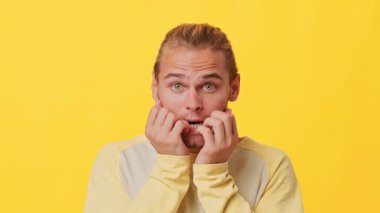 Karışık soydan gelen genç bir adam, parlak sarı bir arkaplanın önünde dururken tırnaklarını yiyerek gergin bir yüz ifadesi sergiliyor. Ruh hali, endişe dolu bir anı yakalıyor..