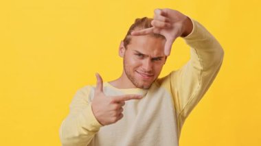 Genç bir adam canlı sarı bir arkaplan önünde, ellerini kullanarak bir çerçeve oluşturmak için şakacı bir şekilde poz veriyor. Kendine güveni yaratıcılık ve enerji hissini yansıtıyor. Z jenerasyonunun tipik özelliği..