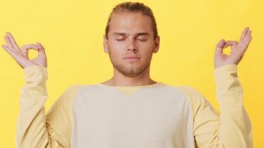 Sakin bir ifadeye sahip genç bir adam, elleri huzurlu bir pozisyonda otururken meditasyon yapar. Açık renk bir süveter giymiş, canlı sarı bir ortamda dinginlik yayıyor..