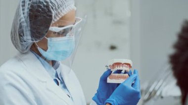 Kişisel koruyucu ekipman kullanan Afrikalı bir dişçi modern bir diş sağlığı kliniğinde diş modelini kullanan bir hastayı diş hijyeni konusunda eğitir..