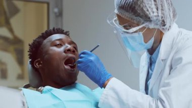 Bir Afrikalı Amerikalı dişçi modern bir diş kliniğinde hastasının dişlerini dikkatle inceliyor. Hasta, dişçi titizlikle ve özenle tedaviye hazırlanırken rahatlamış görünüyor..