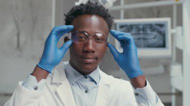 Genç bir Afrikalı Amerikalı dişçi, laboratuvar önlüğü ve güvenlik gözlüğü takarak çağdaş bir diş kliniğinde güvenle duruyor..