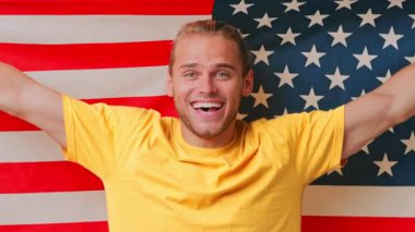 Işıl ışıl gülümseyen genç bir adam, Amerikan bayraklarına karşı coşkuyla poz veriyor. Parlak sarı bir gömlek giyiyor. Şenlikli bir ortamda gurur ve mutluluk simgesi..