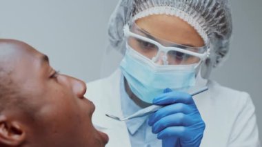 Maske takan ve yüz maskesi takan Afrikalı bir dişçi diş kliniğindeki bir hastayı dikkatle muayene ediyor. Odaklanmış atmosfer diş sağlığı hizmetinin önemini vurguluyor.