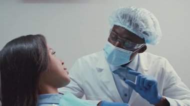 Beyaz önlüklü ve koruyucu kıyafetli bir dişçi, iyi ışıklandırılmış bir diş kliniğinde bir hastayla meşgul oluyor. Hasta diş sağlığı seçeneklerini tartışırken özenli görünüyor..