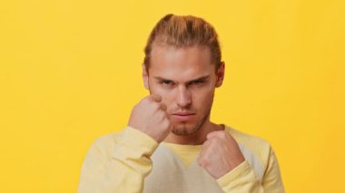 Miras sahibi genç bir adam sert ve kararlı bir ifade sergiliyor. Parlak sarı bir arka plana karşı duruyor, kendine güvenen tavrını ve harekete hazır olduğunu gösteriyor..