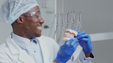 Bir Afrikalı Amerikalı dişçi, çağdaş bir klinikte diş modelini tutarken gülümser. Mankenle takılıyor, diş sağlığı ve hasta eğitimine olan tutkusunu gösteriyor..