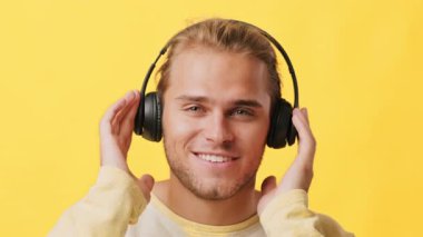 Neşeli bir suratı olan genç bir adam onları kafasına yerleştirirken siyah kulaklık takıyor. Canlı sarı bir arkaplanın önünde duruyor, neşe ve enerji dolu bir anı yakalıyor..