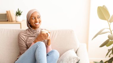 Huzurlu bir hafta sonu. Evde sıcak sıcak bir içecekle kanepeye uzanan tesettürlü pozitif siyahi kadın. Güzel Afrikalı Amerikalı kadın, başörtüsü takmış, kahve molası veriyor, kanepede uzanıyor, fotokopi çekiyor.
