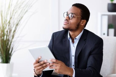 İş dünyasındaki modern teknolojiler. Dalgın Afro-Amerikalı takım elbiseli genç işadamı ofiste dijital tablet kullanıyor, internette çalışıyor, sunuma hazırlanıyor, fotokopi alanına bakıyor.