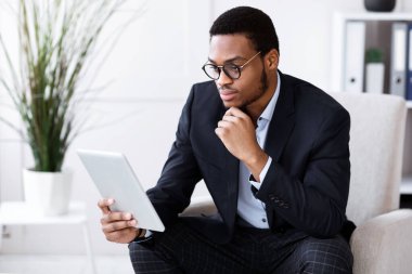 Modern dijital tablet kullanarak, internet üzerinden, ofis içi ve fotokopi alanlarında iş konuşmaları yaparak, gözlüklü siyahi girişimci. Genç Afrikalı Amerikalı yönetici dijital ped tutuyor.