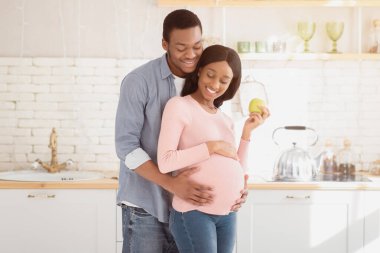 Sevgi dolu zenci bir adamın portresi. Müstakbel karısını mutfakta elma ile kucaklıyor. Mutlu Afrika kökenli Amerikalı çift dengeli hamilelik diyetini destekliyor, bebek beklerken sağlıklı besleniyorlar.