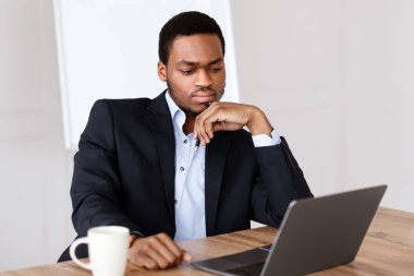 Sorunlu siyah yönetici dizüstü bilgisayara bakıyor, kahve içiyor, işiyle ilgili sorunları var, ofis içi, fotokopi alanı. İş hayatında kriz, iş konseptinde zorluklar