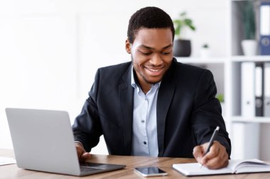 Gülümseyen Afrikalı Amerikalı girişimci ofiste online eğitim alıyor, dizüstü bilgisayar kullanıyor, notlar alıyor, kopyalama alanı kullanıyor. Neşeli siyah işadamı bilgisayarla çalışıyor ve not defterinde yazıyor.
