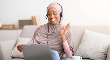 Mutlu siyah Müslüman kadın tesettür ve kulaklık takıp dizüstü bilgisayarından ekranda el sallıyor. Çevrimiçi iletişimden zevk alan sevimli genç Arap kadın web konferansına katılıyor.