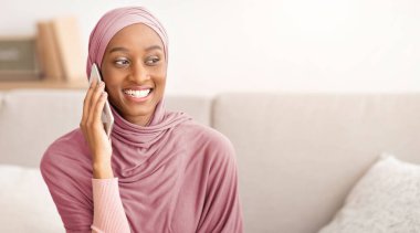 Evde tesettürlü gülümseyen siyah kadın akıllı telefondan konuşuyor. Geleneksel türbanlı pozitif Müslüman kadın cep telefonuyla iletişim kuruyor, ailesiyle ya da arkadaşlarıyla sohbet ediyor.