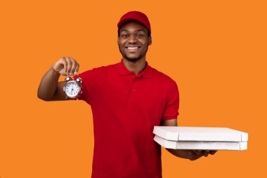 Hızlı Pizza Teslimatı. Kırmızı şapkalı ve üniformalı gülümseyen Afro-Amerikan erkek kuryenin portresi restorandan pizza kutusunu tutuyor ve turuncu sarı stüdyo arka planında izole edilmiş saati gösteriyor.