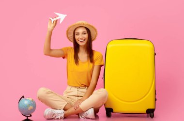 Kağıt uçakla oynayan mutlu bir bayan, bavul ve kürenin yanında oturan, seyahat etmeye hazır, pembe stüdyo geçmişi olan. Yazlık giysiler içindeki mutlu genç kadın uçuştan önce hazırlanıyor.