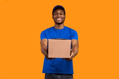 En iyi servis konsepti. Mavi şapkalı ve biçimsiz gülümseyen siyah teslimatçı elinde karton kutuyla, sarı turuncu stüdyo arka planında izole bir şekilde duran kameraya gösteriyor ve veriyor.