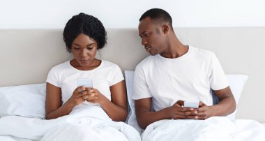 Zenci adam kız arkadaşını cep telefonuyla dinliyor, akıllı telefon ekranına bakıyor. Afrika kökenli Amerikalı genç bir çift yatakta oturuyor ve cep telefonu kullanıyor. İlişkilerde güvensizlik
