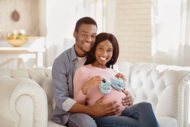 Minik bebek kalçalı zenci hamile bir kadın ve evinde kanepesinde mutlu kocası sarılıyor. Müstakbel anne ve baba, bebek bekleyen küçük ayakkabılarla, kanepede kucaklaşıyorlar.