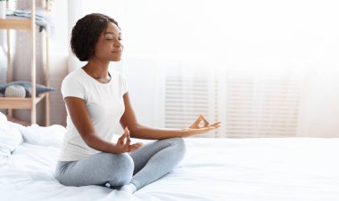 Nilüfer pozisyonunda yatağında oturmuş, meditasyon yapan, gözleri kapalı, rahat siyah kadın, kopyalama alanı. Sağlıklı yaşam tarzı kavramı. Güzel Afrikalı Amerikalı genç bayan yatak odasında sabah yogası yapıyor.