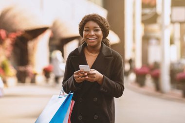 Kara Cuma 'da mobil uygulama ve nakde çevirme ve sezon satışı konsepti. Akıllı telefonlu mutlu Afro-Amerikan bayanı alışveriş merkezinde, açık havada, serbest alanda başarılı bir alışveriş yaptıktan sonra banka hesabını kontrol ediyor.
