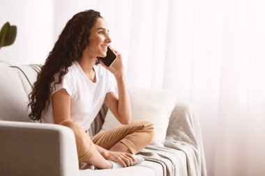 Oturma odasındaki kanepede oturan, cep telefonuyla konuşan, fotokopi odasına bakan güzel genç bir kadın. Koyu kıvırcık saçlı neşeli bayan akıllı telefonda konuşuyor, hafta sonunu evde geçiriyor.