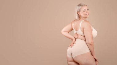 Vücut pozitiftir. Güzel kıvrımlı kadın bej renkli stüdyo arka planında poz veriyor, çekici artı iç çamaşırlı sarışın yarı dönüşmüş ve kameraya gülümsüyor, boşluğu kopyalıyor.