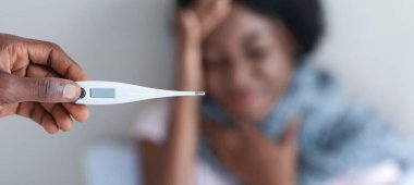 Soğuk, baca, koronavirüs konsepti. Ateşli siyah bir kadın, erkek elinin termometresine odaklanmış. Afro-amerikan bayanın bacası var, başı ağrıyor, kocası ona bakıyor.