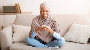 Hoş, tesettürlü siyahi bir kadın akıllı telefonuyla evde rahat bir kanepede otururken mesaj atıyor. Güzel Müslüman bir kadın arkadaşına mesaj atıyor, e-postaları kontrol ediyor, kapalı mekanda cep telefonundan video izliyor.