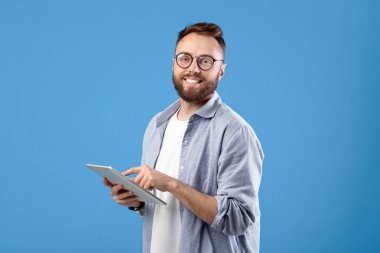 Sıradan giyinen genç, mavi stüdyo arka planında tablet bilgisayar kullanıyor. Dokunmatik bloknotu olan mutlu bir milenyum adamı çevrimiçi çalışıyor, e-posta gönderiyor, video izliyor.