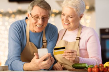 Senior Spouses Shopping Online With Smartphone In Kitchen, Focus On Credit Card And Mobile Phone. Modern Grocery Shopping For Older People Concept. Shallow Depth