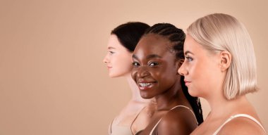 Farklı görünümlü, bej arka planda sutyenlerle poz veren üç güzel vücut portresi, kameraya gülümseyen genç Afrikalı Amerikalı kadın,