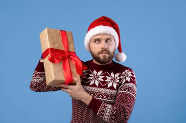 Noel kıyafetli ve Noel Baba şapkalı genç adam mavi stüdyo arka planında hediye kutusunun içinde ne olduğunu dinliyor. Yılbaşı hediyeli milenyum adamı kış tatiline hazır.